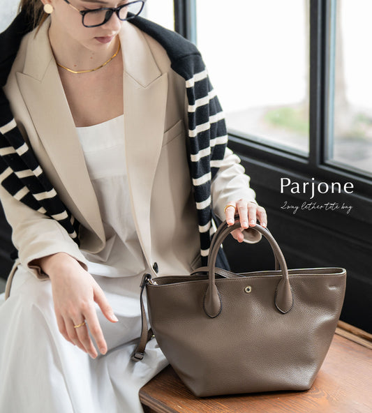 A female model is sitting and holding a tote bag "Parjone". Color:Taupe