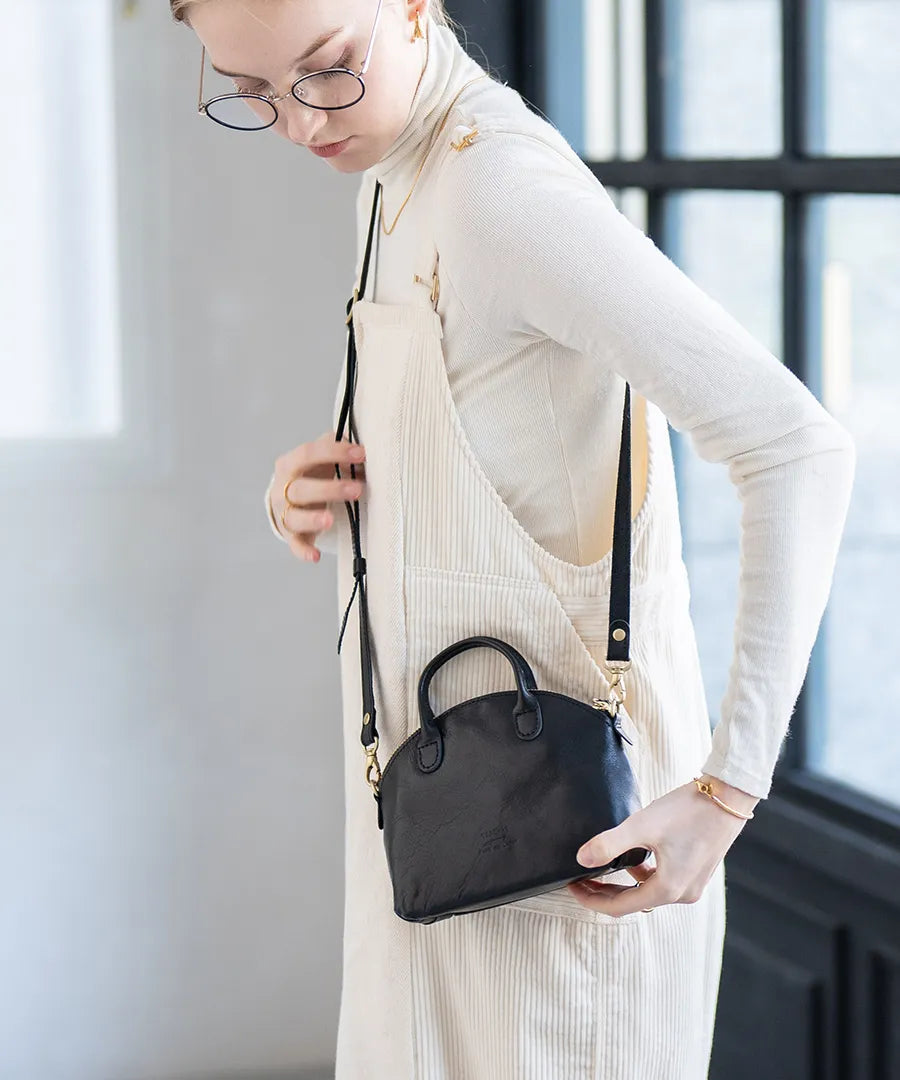 A female model is carrying a bag "Light leather boston mini" over her shoulder. Color:Black