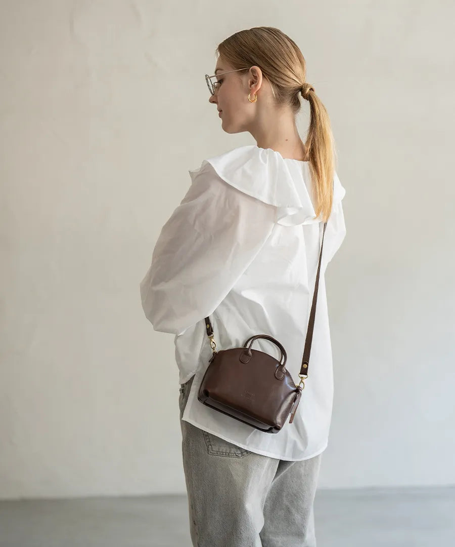 A female model is carrying a bag "Light leather boston mini" over her shoulder. Color:Chocolate brown