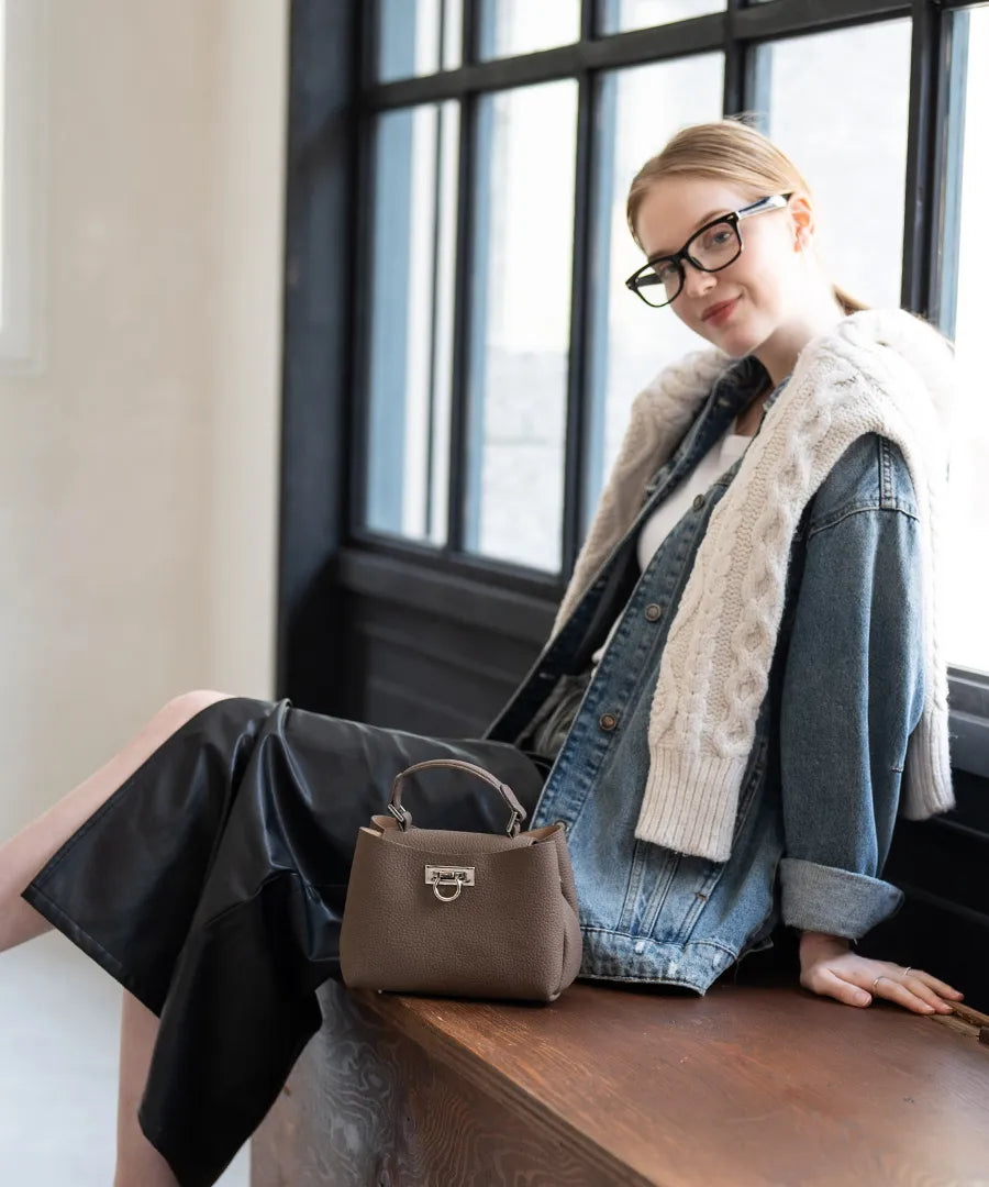 A female model is sitting and holding a tote bag "Baby Marollet". Color: Taupe