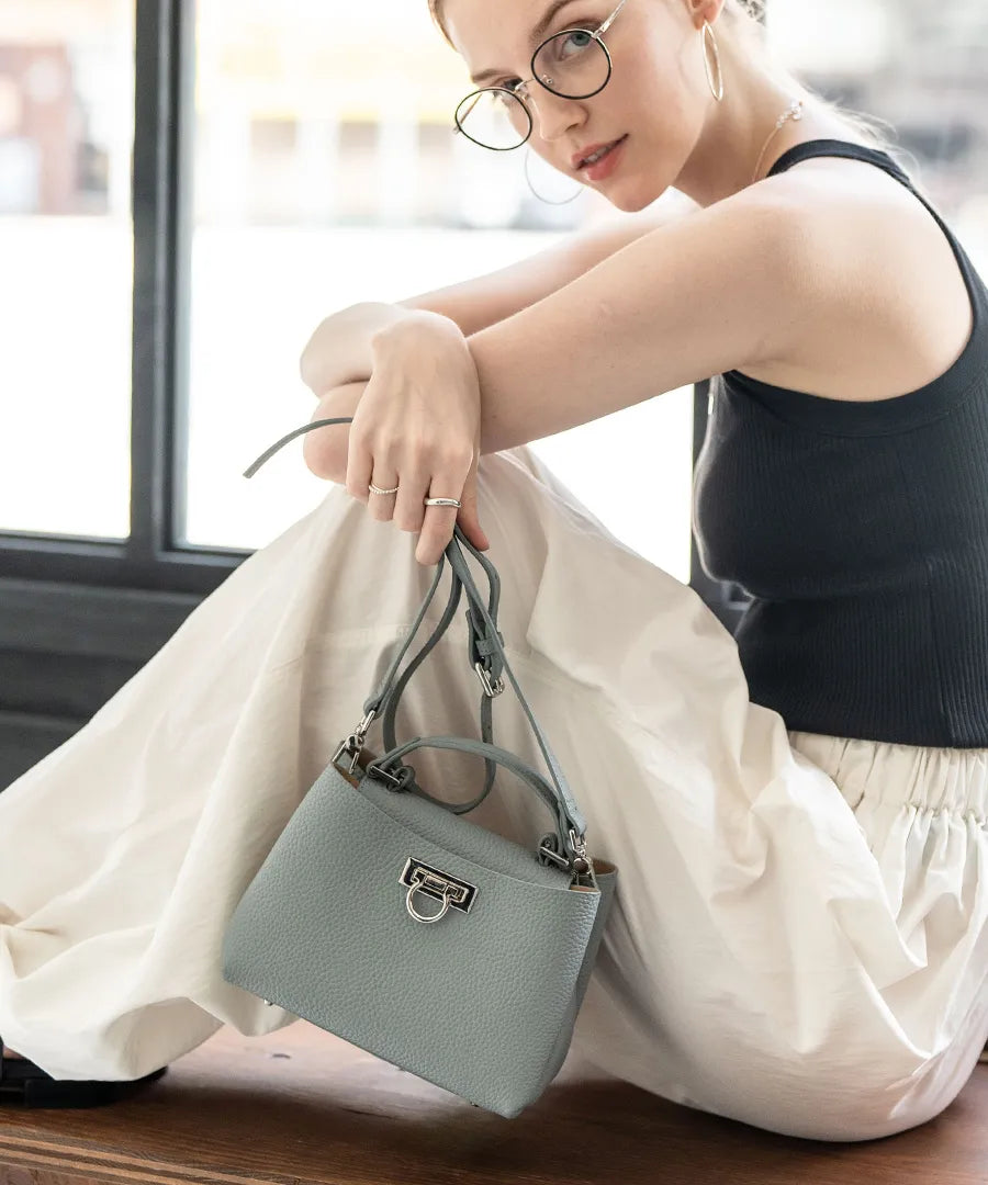 A female model is sitting and holding a tote bag "Baby Marollet". Color:Blue Gray