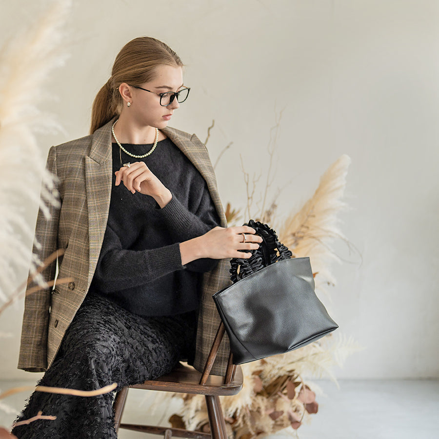 A female model is sitting and holding a hand bag "Frillka". Color:Black x Black