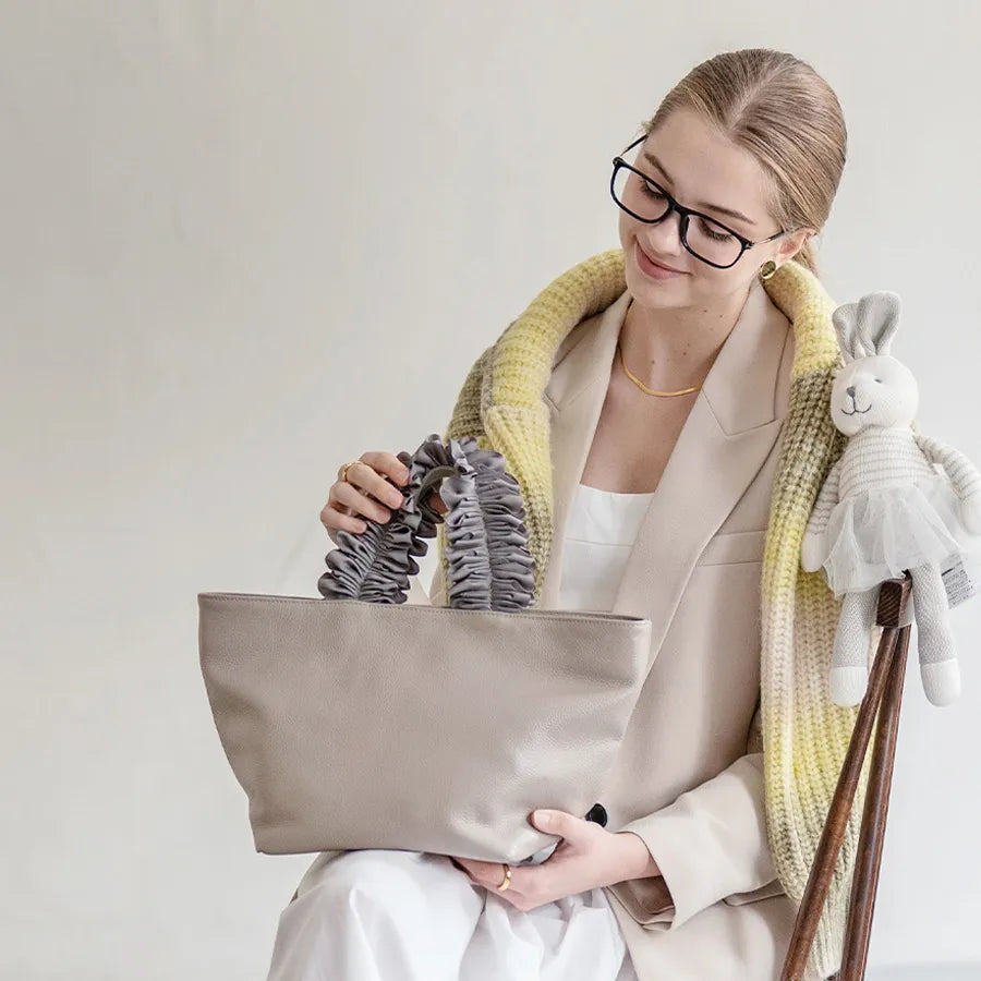 A female model is sitting and holding a hand bag "Frillka". Color:Light greige x Gray