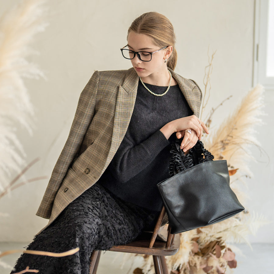 A female model is sitting and holding a hand bag "Frillka". Color: Black x Black