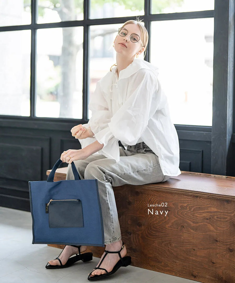 A female model is sitting and holding a tote bag "lesche". Color: Navy