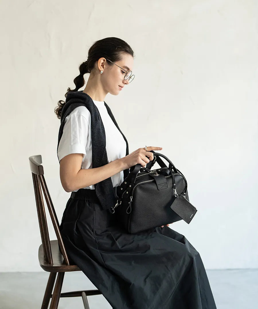 A female model is sitting and holding a hand bag "Mini Loche (Version 7)".Color:Black