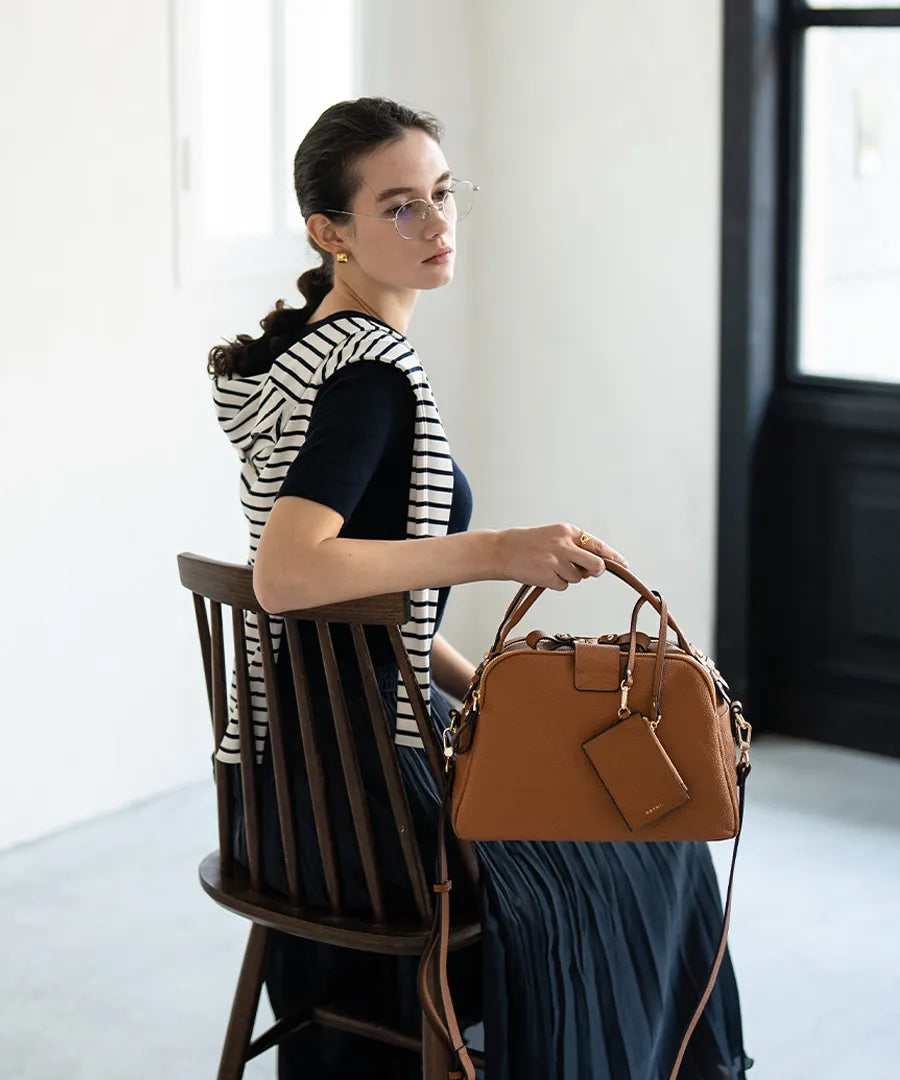 A female model is sitting and holding a hand bag "Mini Loche (Version 7)".Color:Camel