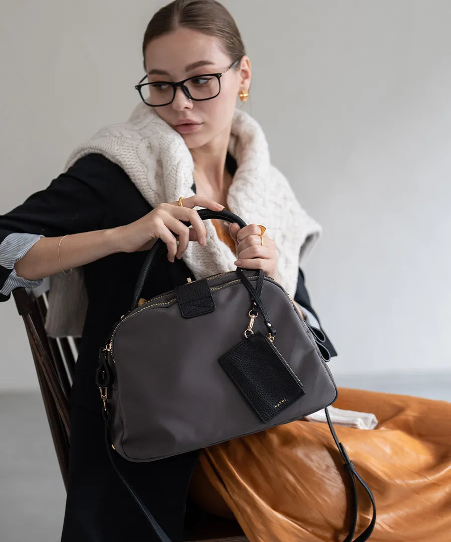 A female model is sitting and holding a hand bag "Mini Nylon Loche".Color:Gray Black