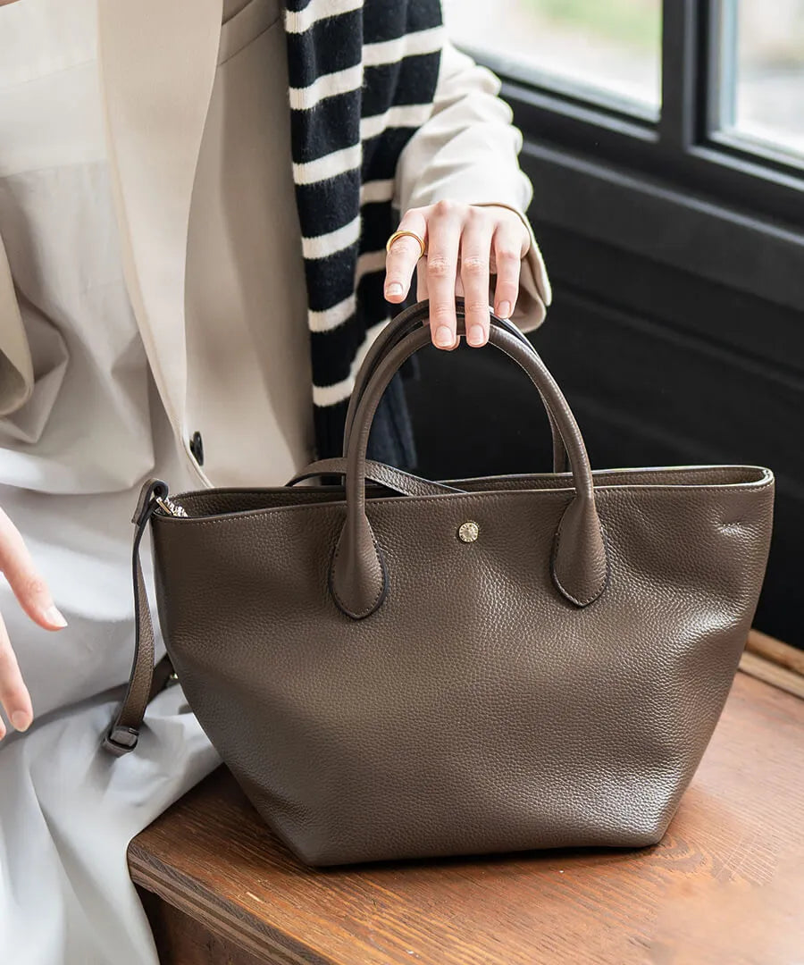 A female model is sitting and holding a tote bag "Parjone". Color:Taupe